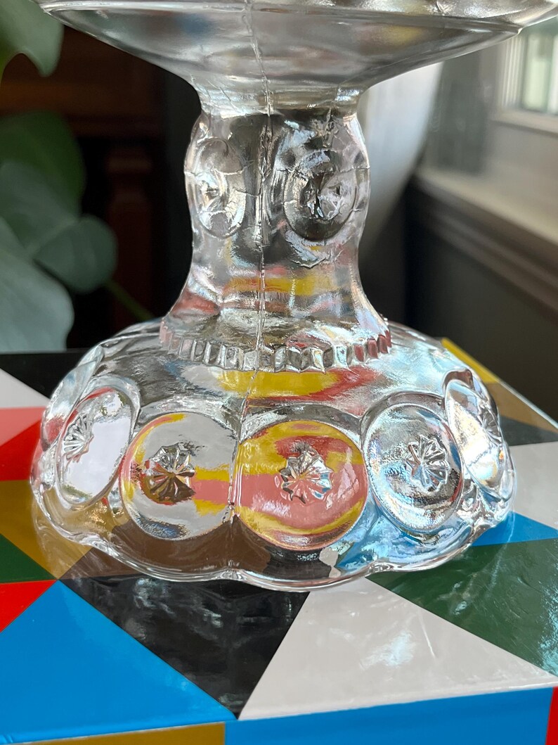 Op de afbeelding: Een glazen voetstuk voor een taart met een gegolfde rand en een voet met een herhalend patroon van cirkels. De voet heeft een getextureerd oppervlak met een subtiele iriserende glans.