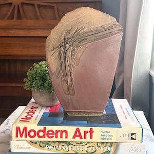 May include: A sculptural ceramic vase with a matte mauve finish and a textured, raised gold-toned relief pattern on the upper section. The base of the vase features a signature and the year 1994 etched into the clay surface.