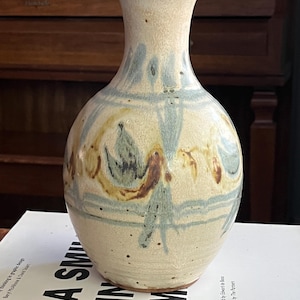 Puede incluir: Un jarrón de cerámica de color crema con la parte superior acampanada. El jarrón presenta diseños abstractos pintados en azul, marrón y amarillo. El jarrón está sobre un libro.