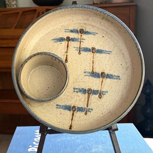 Può includere: Un piatto da portata in ceramica con una ciotola per salse integrata. Il piatto beige presenta disegni di libellule blu e un bordo blu. È esposto su un piccolo cavalletto in metallo scuro. Il piatto è un pezzo di ceramica unico.