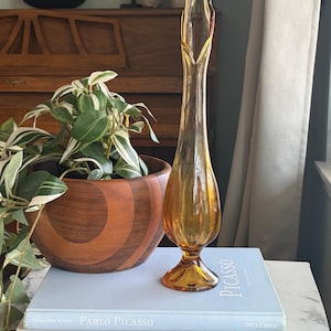 Op de afbeelding: Een hoge, amberkleurige glazen vaas met een uitlopende voet en een lange hals. De vaas staat op een stapel boeken, waaronder een met de titel "Picasso". Een houten kom met een plant is op de achtergrond te zien.