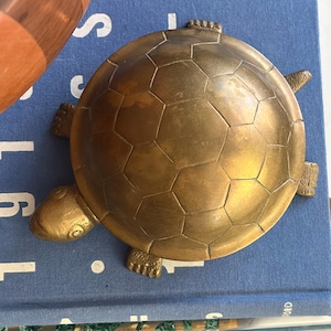 May include: A brass turtle figurine with a detailed hexagonal shell pattern, a smooth rounded back, and four textured feet. The turtle has a small head with visible eye markings and is positioned with its limbs extended outward from the shell.