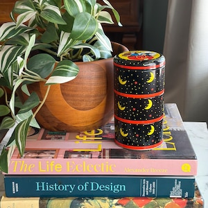 May include: A black and red decorative tin with a celestial design of moons and stars, stacked on top of books. A potted plant with green and white leaves sits to the left. The books include titles like "The Life Eclectic" and "History of Design."