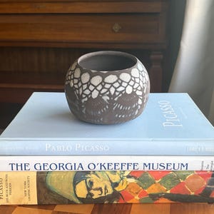 May include: A brown ceramic bowl with a white lace-like pattern sits atop a stack of books. The books include titles like "Pablo Picasso" and "The Georgia O'Keeffe Museum". The bowl has a round shape and a dark interior.