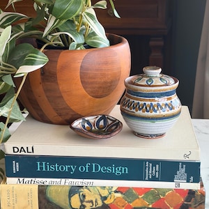 May include: A wooden bowl with a plant, a small ceramic dish, and a ceramic jar with a lid sit atop a stack of books. The books include titles like "History of Design" and "Matisse and Fauvism."