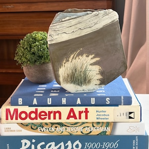May include: A flat, sculptural ceramic vase with a textured, off-white top section and a smooth, dark gray lower body featuring a hand-painted illustration of green beach grass. The piece is signed by the artist and dated 90 at the base.