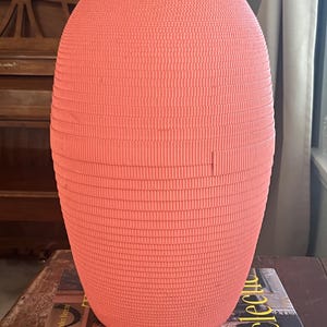 May include: A coral pink ceramic vase with a textured, ribbed surface. The vase is oval shaped and has a wide opening at the top.