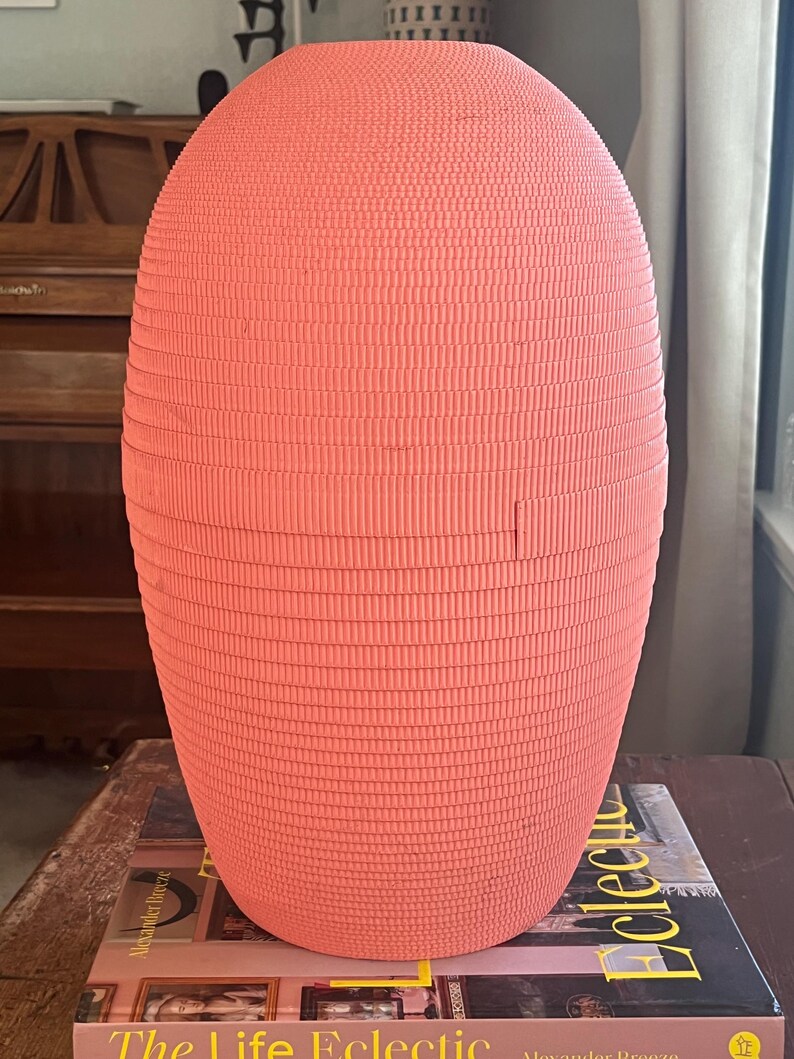 May include: A coral pink ceramic vase with a textured, ribbed surface. The vase is oval shaped and has a wide opening at the top.