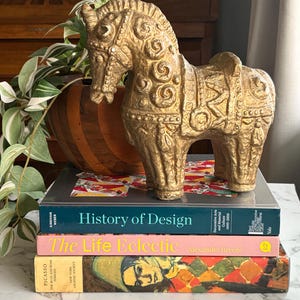 Puede incluir: Una figura decorativa dorada con forma de caballo con detalles intrincados, colocada sobre una pila de libros. Los libros incluyen "History of Design" y "The Life Eclectic". La figura es un objeto decorativo.