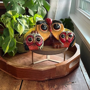 May include: Three hand-painted wooden owls, one large and two smaller, are arranged on a wooden tray with a metal stand. The owls are painted in shades of brown, yellow, red, and black with large, expressive eyes.