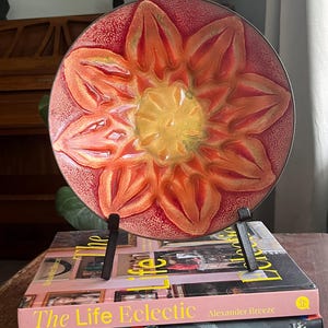 May include: A red and orange ceramic plate with a flower design. The plate is on a black metal stand and is sitting on top of two books. The top book is titled "The Life Eclectic" by Alexander Breeze. The bottom book is titled "modernstarts PEOPLE PLACES THINGS".