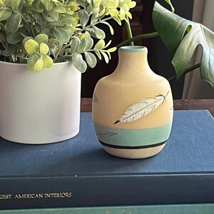 Może przedstawiać: Mały, jasnożółty ceramiczny wazon z wzorem niebieskiej i białej pióra. Wazon stoi na dwóch niebieskich i zielonych książkach.