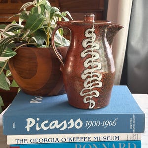 May include: A ceramic pitcher with a lid, featuring a reddish-brown glaze and a white, leaf-like design. The pitcher sits atop a stack of books, including one titled "Picasso 1900-1906". A wooden planter with a green plant is in the background.