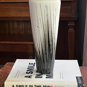 Può includere: Vaso alto, bianco sporco, con bordo nero e un disegno di linee nere verticali. Il vaso è esposto su una pila di libri, tra cui uno intitolato "A Smile in the Mind".