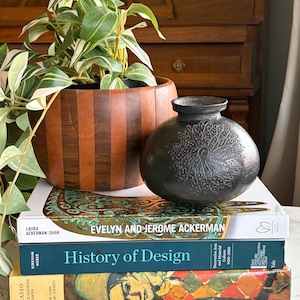 Könnte beinhalten: Ein Stillleben mit einer dunkelgrauen Vase mit floralem Muster, einem Holzpflanzgefäß mit einer grünen Pflanze und einem Stapel Bücher. Die Bücher tragen Titel wie "Evelyn and Jerome Ackerman" und "History of Design."