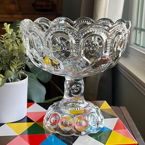 Op de afbeelding: Een glazen schaal op een voet met een gegolfde rand en een getextureerd, verhoogd patroon van cirkels. De schaal staat op een kleurrijk geometrisch oppervlak.