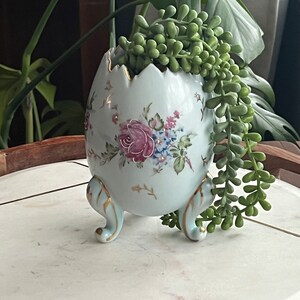 May include: A white ceramic egg-shaped planter with gold accents and floral designs. The planter has three gold feet and is filled with a green succulent plant.
