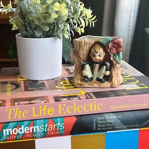 Könnte beinhalten: Ein Keramik-Pflanztopf in Form eines Baumstamms mit einer kleinen Gnomenfigur, die herausguckt. Der Gnom trägt einen braunen Hut, eine grüne Jacke und eine braune Hose. Der Topf hat eine rote Blume und grüne Blätter an der Seite. Der Topf steht auf einem Stapel Bücher mit den Titeln "The Life Eclectic" und "modernstarts".