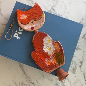 May include: A ceramic cat-shaped wind chime featuring a stylized orange face with closed eyes and a red nose, connected to a larger orange body decorated with white flowers and green leaves, finished with a small terracotta bell at the bottom.