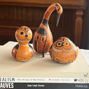 Può includere: Tre statuette di gufo in legno fatte a mano di varie dimensioni e forme. I gufi sono principalmente arancioni con dettagli marrone scuro. È presente anche una statuetta di uccello alta e sottile con un lungo collo e un becco. Le statuette sono esposte sopra i libri.