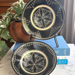 Puede incluir: Dos platos decorativos negros con un diseño geométrico crema. Los platos se exhiben sobre un soporte de metal, con una planta y libros en el fondo. Los platos tienen un acabado brillante y un aspecto artístico único.