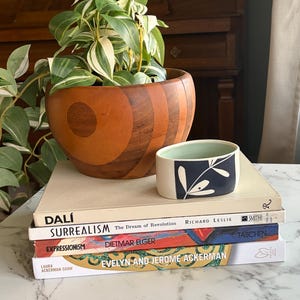 May include: A wooden bowl with a plant sits atop a stack of books, including titles on Surrealism and Expressionism. A small ceramic container with a dark blue and white leaf design is also present. The scene is set on a marble surface.