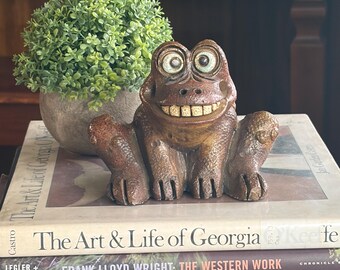 May include: A whimsical, brown frog figurine with large, expressive eyes and a toothy grin. The frog is perched atop a stack of books, including titles like "The Art & Life of Georgia O'Keeffe." A small, green plant in a pot sits nearby.