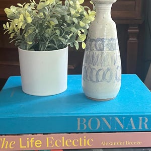 以下が含まれることがあります： 青い抽象的なデザインの白いセラミック花瓶が、「BONNAR」というタイトルの青い本の上に立っています。左側には、緑と黄色の葉が入った白い植木鉢があります。「The Life Eclectic」というタイトルの本など、他の本も見えます。