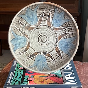 Może przedstawiać: Ceramiczna miska z kwiatowym wzorem w odcieniach niebieskiego i brązowego. Misa spoczywa na stosie książek z tytułami takimi jak "Matisse" i "Fauves". Wnętrze miski ma spiralny wzór.