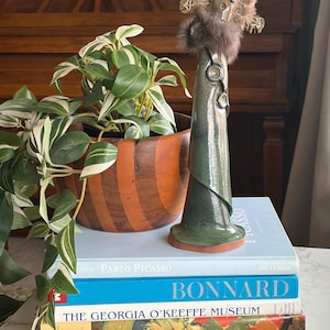 Puede incluir: Un jarrón decorativo verde y plateado, adornado con piel y joyas. Descansa sobre una pila de libros de arte. Una planta en maceta con hojas verdes y blancas está en el fondo. Los libros incluyen títulos sobre Picasso y Bonnard.
