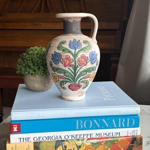 May include: A vintage ceramic vase with a handle, featuring a floral design in blue, red, and yellow. The vase sits atop a stack of books, including titles like "Pablo Picasso" and "Bonnard", creating a decorative display.