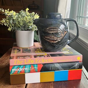 Może przedstawiać: Niebiesko-brązowy ceramiczny czajnik z motywem ryb, stojący na dwóch książkach. Górna książka nosi tytuł "The Life Eclectic", a dolna "modernstarts PEOPLE PLACES THINGS".