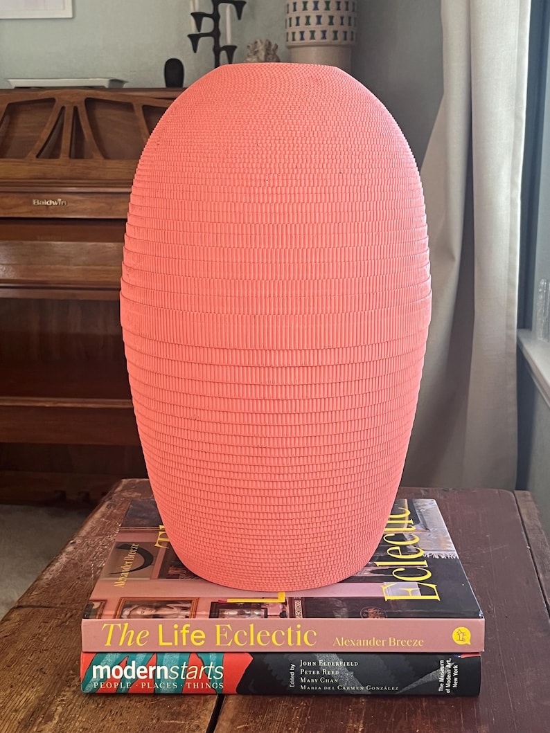 May include: A coral pink ceramic vase with a ribbed texture sits on top of two books. The top book is titled "The Life Eclectic" by Alexander Breeze. The bottom book is titled "modernstarts PEOPLE PLACES THINGS".