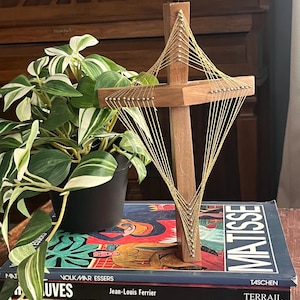 Puede incluir: Una cruz de madera con detalles de hilo dorado, de pie sobre una pila de libros de arte. La cruz tiene un poste vertical y una viga horizontal, con el hilo dorado creando un diseño geométrico. Los libros incluyen el título "MATISSE". Una planta en maceta está en el fondo.