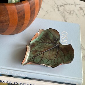 Peut inclure: Un plat décoratif en forme de feuille avec une patine verte et marron. Le plat repose sur une pile de livres, dont un intitulé "Pablo Picasso". Le plat est probablement en métal et est un objet décoratif.