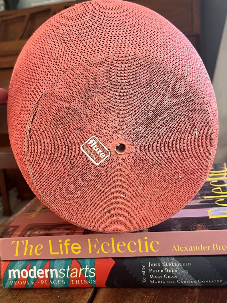 May include: A red, round, cardboard object with a textured surface. The object has a small hole in the center and a white label with the word "flute" printed on it. The object is sitting on top of two books, one titled "The Life Eclectic" and the other titled "modernstarts PEOPLE PLACES THINGS".