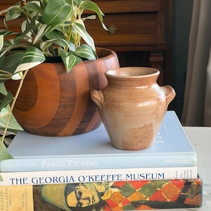 May include: A small, tan ceramic vase with two handles rests on a stack of books. A wooden planter with a plant is behind the vase. The books include titles such as "Pablo Picasso" and "The Georgia O'Keeffe Museum."