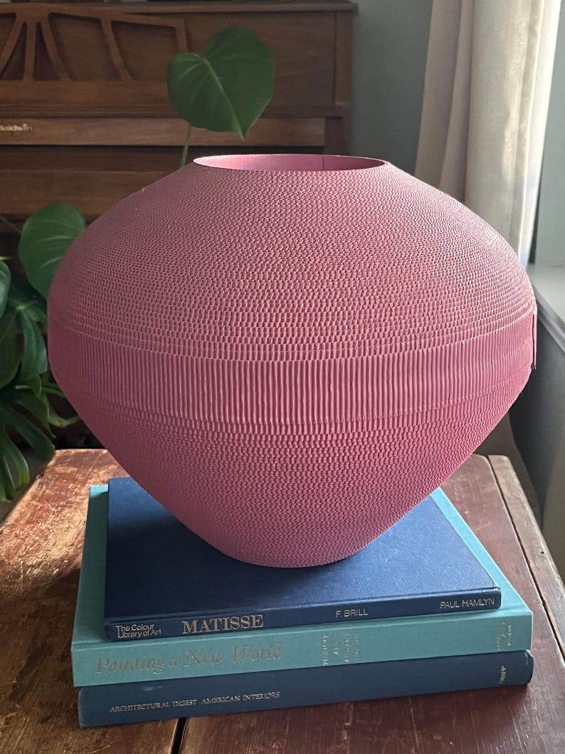 May include: A pink, corrugated cardboard vase sits on top of a stack of three books. The books are blue and teal with titles that include "Matisse" and "Painting a New World".