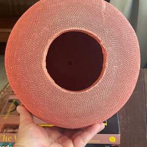 May include: A coral-colored, round, woven paper lampshade with a large circular opening in the center. The lampshade is made of a delicate, intricate pattern of woven paper.