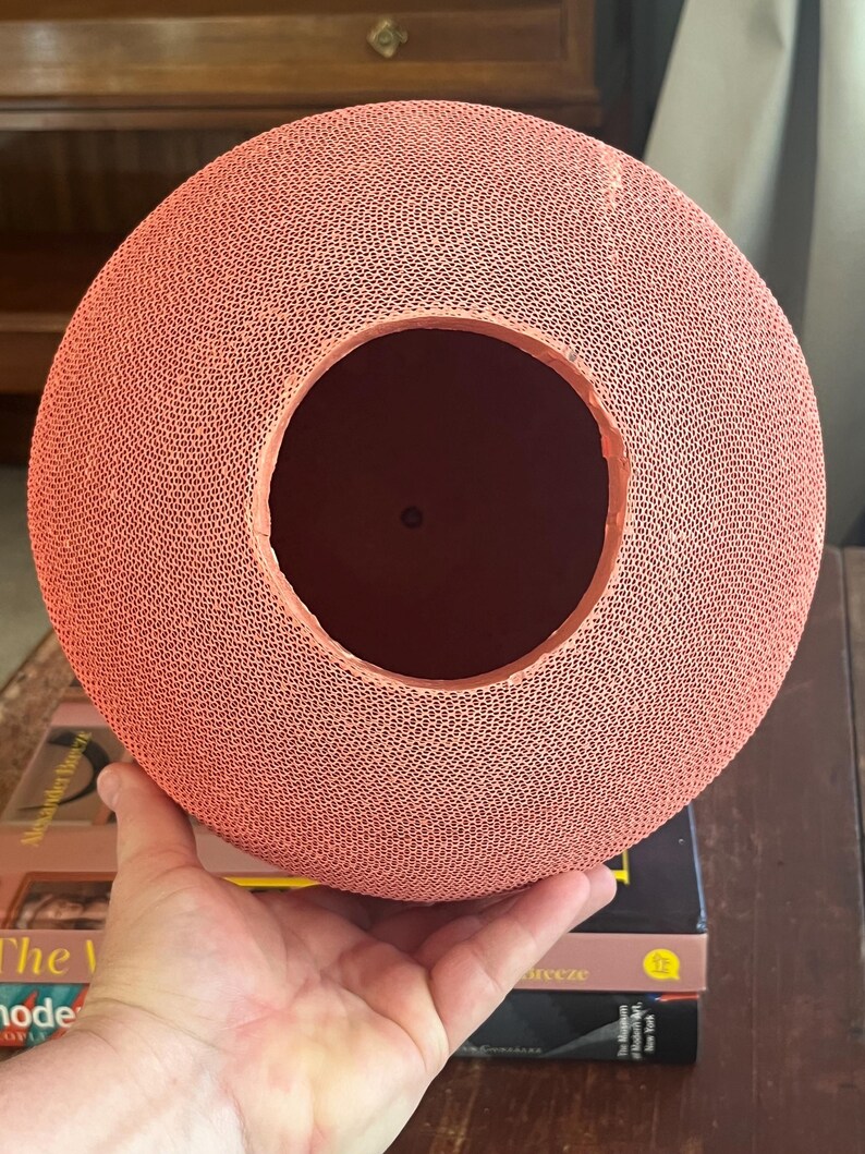 May include: A coral-colored, round, woven paper lampshade with a large circular opening in the center. The lampshade is made of a delicate, intricate pattern of woven paper.