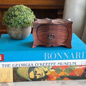 May include: A small, intricately carved wooden box with a drawer and a metal handle. The box is a rich reddish-brown color and sits atop a stack of books, including one titled "BONNARD". A small, green plant in a gray pot is also visible.