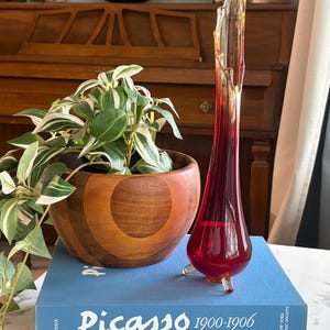 May include: A slender, red glass vase with a flared top and three small feet. It rests on a stack of books, including one titled "Picasso 1900-1906." A wooden bowl with a plant is next to the vase. The background includes a wooden piano.