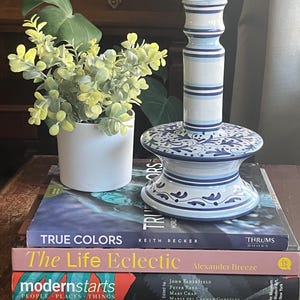 May include: A blue and white ceramic candle holder with a striped design. The candle holder is sitting on a stack of three books. The top book is titled "The Life Eclectic" by Alexander Breeze. The middle book is titled "True Colors" by Keith Recker. The bottom book is titled "modernstarts: People Places Things".