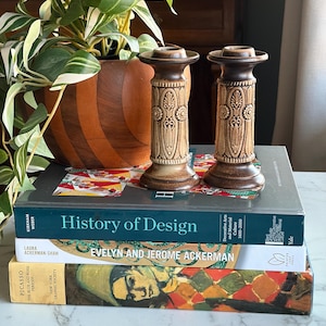 Op de afbeelding: Twee bruine en beige kandelaars met sierlijke bloemmotieven staan op een stapel boeken. De boeken bevatten titels als "History of Design" en "Picasso". Een potplant met groen-witte bladeren is zichtbaar op de achtergrond.