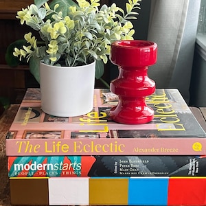 Puede incluir: Una pila de tres libros con los títulos "The Life Eclectic", "modernstarts PEOPLE PLACES THINGS" y "The Museum of Modern Art, New York". Un portavelas de cerámica rojo se encuentra encima de los libros.