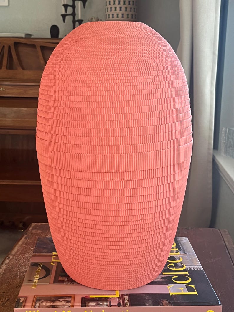 May include: A large, pink, ceramic vase with a textured, ribbed surface. The vase is oval shaped and has a smooth, rounded top.