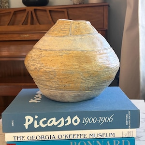 May include: A textured, yellow and beige ceramic vase with a rounded body and a narrow neck. The vase sits atop a stack of books, including one titled "Picasso 1900-1906". The books are various colours, including blue and white.