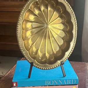 May include: A decorative, gold-colored brass tray with a scalloped design and beaded edge. The tray is displayed on a black metal stand, resting on a stack of books with titles including "BONNARD" and "The Life Eclectic".
