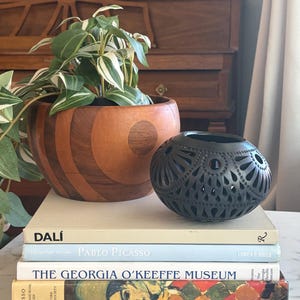 Puede incluir: Una maceta de madera con una planta verde y blanca se encuentra junto a una esfera negra decorativa calada. Libros apilados con títulos como "Dali", "Pablo Picasso" y "The Georgia O'Keeffe Museum". Los libros están sobre una superficie de mármol.