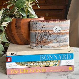 Puede incluir: Un jarrón rectangular de cerámica con un diseño de paisaje con una luna y ramas, sobre una pila de libros. A la izquierda, una maceta de madera con una planta verde y blanca.